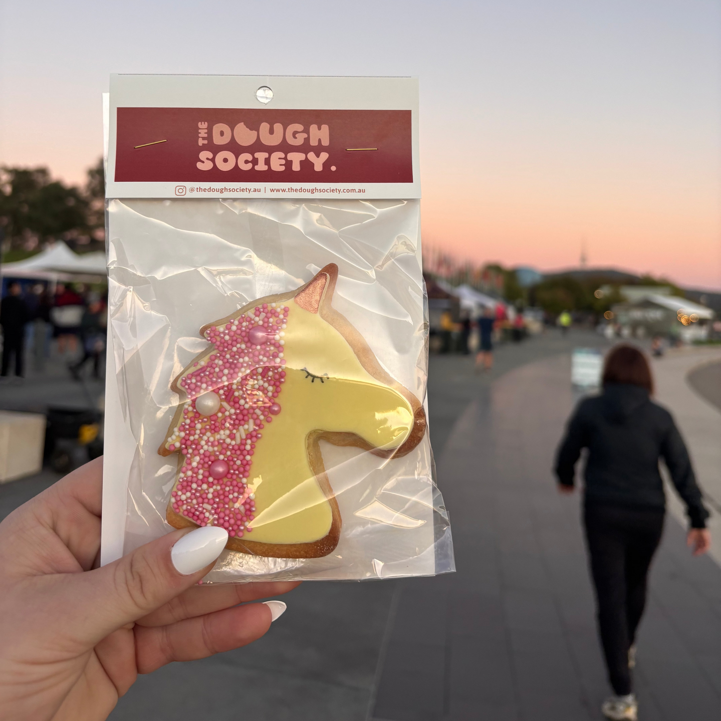 Pastel yellow fondant unicorn cookie with white and pink sprinkles being held in the foreground on the bottom left of photo. The Canberra sunrise in the background and the Telstra Tower is just visible although blurry.