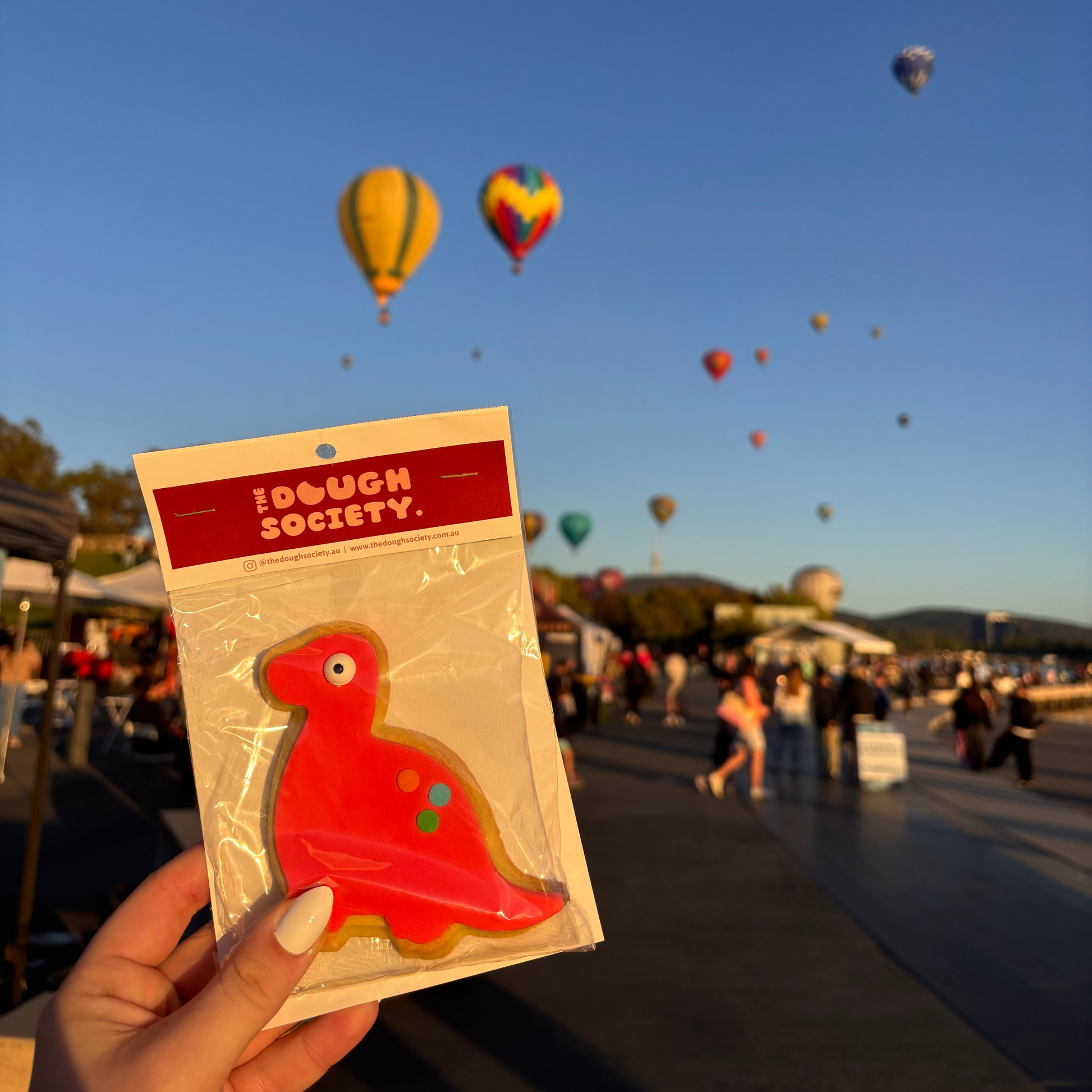 Red dinosaur-shaped product with 'The Dough Society' branding held in front of a hot air balloon festival.